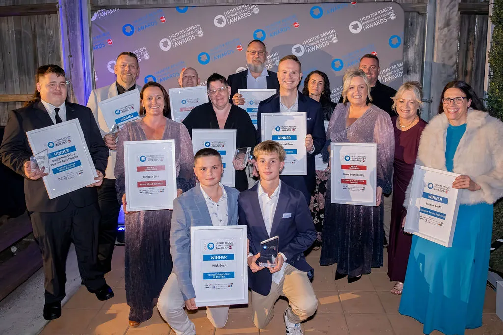 A group of award recipients in formal dress holding framed certificates and glass trophies in front of a Hunter Region Business Excellence Awards backdrop